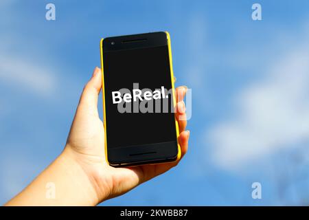 Hand holding smartphone in yellow plastic case on blue sky nature background. Bereal app. BeReal is a new social media app and Photo sharing platform. Stock Photo