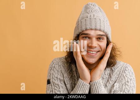 Young hadsome smiling man with long hair in winter hat holding his cheeks over isolated brown background Stock Photo