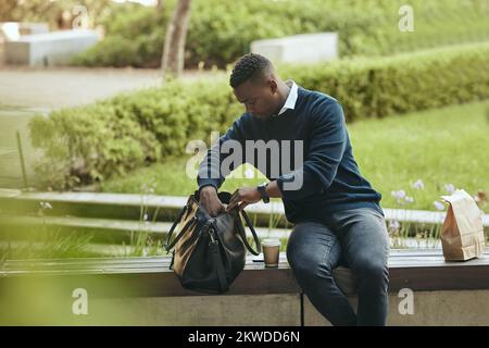 Businessman city park, food and lunch break with coffee or tea drink in paperbag. Young African entrepreneur looking in bag for strategy notebook and Stock Photo