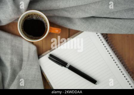A orange Cup of hot coffee with a notebook on wooden table with black pen. Autumn mood concept. Winter mood concept. Warm autumn or winter picture. Stock Photo