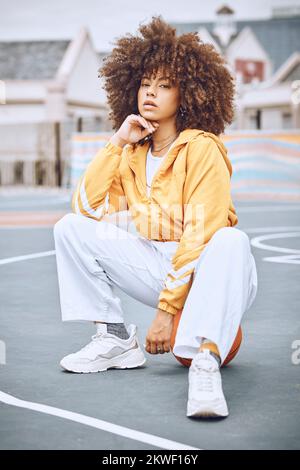 Trendy, stylish and fashionable female basketball player sitting on a ball at the court against an urban background. Young, edgy and serious woman Stock Photo
