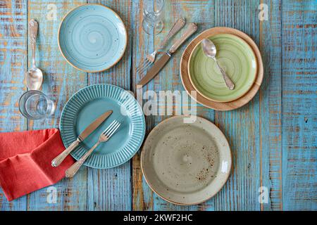 Set of porcelain plates of different types and colors on a blue planked table with crystal glasses and cutlery on the plates Stock Photo