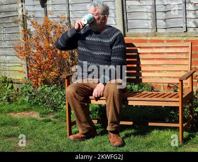 Elderly or senior man sitting on a bench drinking a cup of tea or coffee. Enjoying his drink in the sunshine. Stock Photo