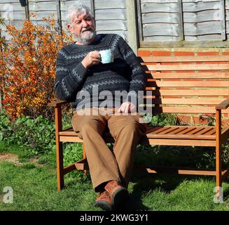Elderly or senior man sitting on a bench drinking a cup of tea or coffee. Enjoying his drink in the sunshine. Stock Photo