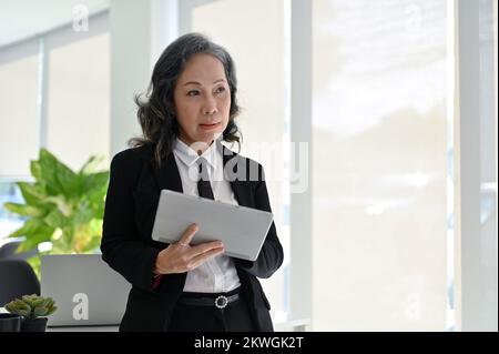 Professional Asian aged businesswoman or female boss in formal business suit using digital tablet, leaning on table, working in her office. Stock Photo