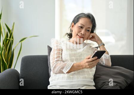 Charming and relaxed Asian retire woman using her smartphone while sitting on sofa in her living room. Stock Photo