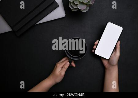 Modern stylish black workspace tabletop with a female hands holding a cup of coffee and smartphone white screen mockup. top view Stock Photo