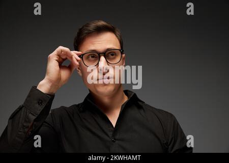 Serious young man, manager in black business classic style shirt putting eyewear on isolated over grey background. Fashion look of businessman. Office Stock Photo