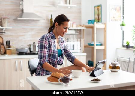 Woman browsing on tablet pc during breakfast in kitchen and holding cup of green tea. Working from home using device with internet technology, typing, on gadget during breakfast. Stock Photo
