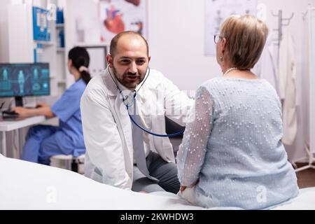 Doctor using stethoscope during consultation of senior woman in hospital. Medical practitioner examining lungs of caucasian in modern clinic dressed in white coat. Stock Photo