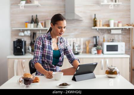 Cheerful woman smiling and reading news on tablet while drinking green tea. Working from home using device with internet technology, typing, on gadget during breakfast. Stock Photo