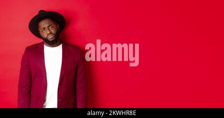 Sad and unamused Black man looking with pity at camera, frowning disappointed, standing against red background Stock Photo