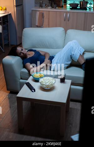 Young woman in pijamas resting lying on couch in front tv smiling. Happy, amused, lonely lady enjoying the evening sitting on comfortable sofa watching television eating popcorn and snacks Stock Photo