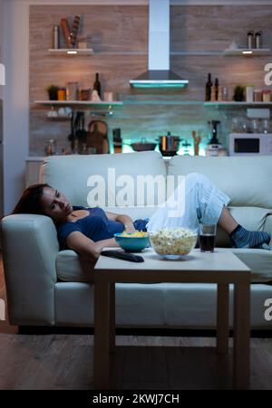 Exhausted woman watching tv show lying on sofa in living room. Tired lonely unhappy young lady in pijamas relaxing on comfortable couch in front of television feeling bored eating snacks late at night Stock Photo