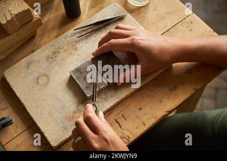 Top view closeup of artisan jeweler holding handmade ring with tweezers over rustic wooden table Stock Photo