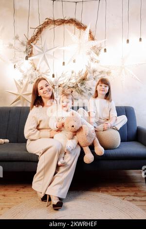 Loving family with mom and two adorable daughters relaxing on the sofa in decorated living room, celebrating Christmas and New Year holidays. Looking Stock Photo