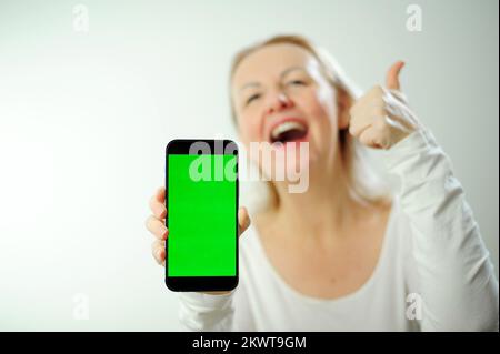 pretty sweet woman in white hoodie looks at the phone, she is glad that she sees there and shows the phone with green screen to camera. Happy lady shares information with us thumbs up show class  Stock Photo