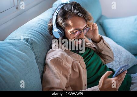 Smiling young man wearing headphones listen music playing in smartphone sitting on sofa at home Stock Photo