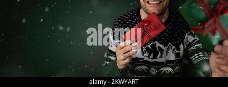 smiling man in christmas sweater showing wrapped gift boxes on green background. banner with copy space Stock Photo