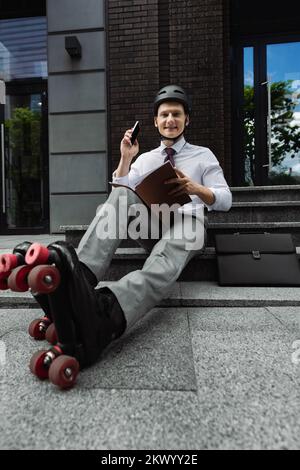 cheerful man in roller skates and formal wear looking at camera while sitting on stairs with smartphone and folder,stock image Stock Photo