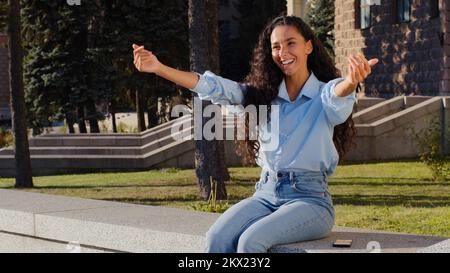 Young beautiful brunette girl woman smiling waiting sitting in city street looking at mobile phone reading message waving hello greeting gesture Stock Photo