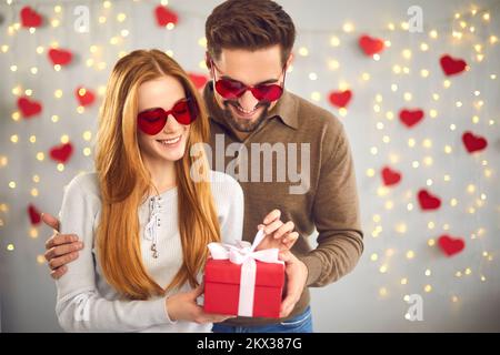 Loving couple unpacks a gift for Valentine's Day standing on a background of garlands and hearts. Stock Photo