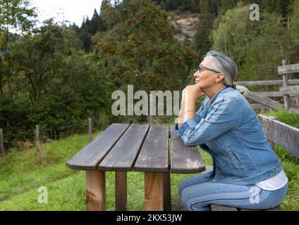 A gray-haired woman sits at a wooden table in the countryside at the edge of the forest with a kind look. Stock Photo