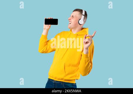 Portrait of overjoyed caucasian guy singing song and using cellphone as microphone, wearing wireless headset Stock Photo