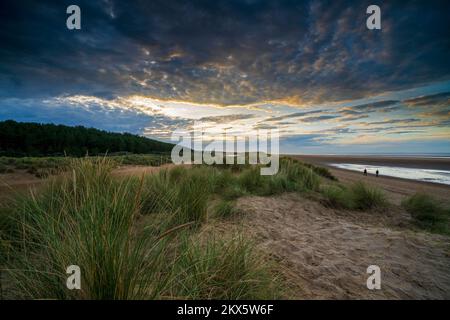 A couple walk along the beach and sand dunes during sunset at Holkham beach, Norfolk, England, Uk. Stock Photo