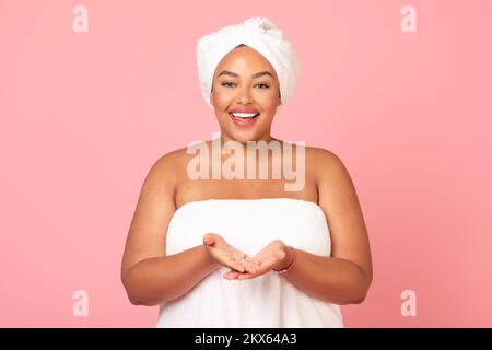 Happy black obese lady wrapped in bath towel demonstrating something on empty palms, showing invisible object Stock Photo