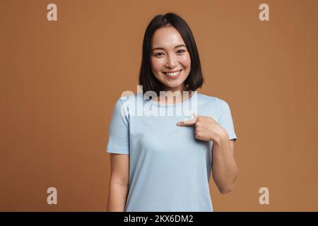 Young asian woman laughing and pointing finger at herself isolated over beige background Stock Photo