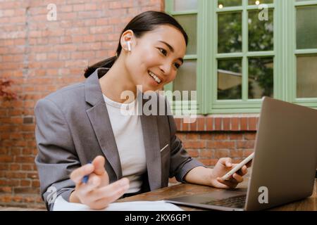 Asian woman in earphones using gadgets while working at cafe outdoors Stock Photo