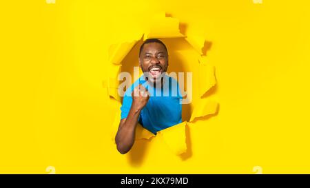 Overjoyed african american man raising clenched fist and shouting, breaking through yellow paper background, panorama Stock Photo