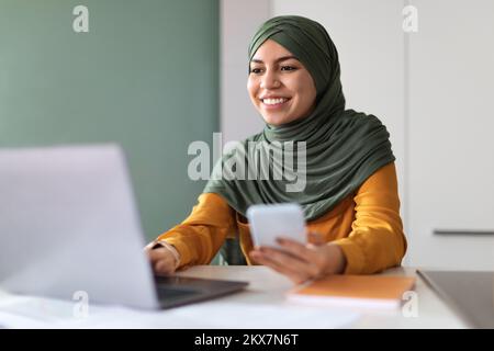 Arab Woman In Hijab Using Smartphone And Laptop While Working At Office Stock Photo