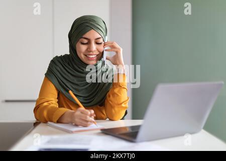 Smiling Muslim Woman In Hijab Talking On Cellphone And Noting Down Information Stock Photo