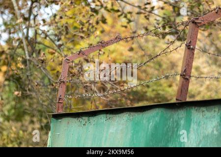Green metal rusty fence with barbed wire on top with greenery, tree branches background. Territory border protection Stock Photo