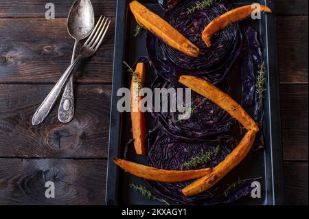 Roasted red cabbage with sweet potatoes on a baking sheet isolated on dark wooden background. Flat lay Stock Photo