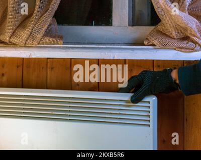 a hand in glove setting temperature on electric heater, hanging under window, indoor close up Stock Photo