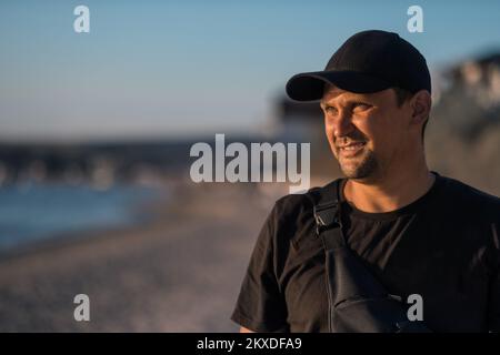 A handsome brunette man in a black t-shirt smiles and looks at the camera against the backdrop of mountains and a blue sky. High quality photo. Stock Photo