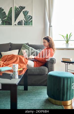 Relaxed young woman sitting on sofa using laptop at home cozy interior. Stock Photo