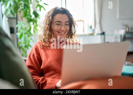 Relaxed happy young woman sitting on sofa using laptop at home surfing. Stock Photo