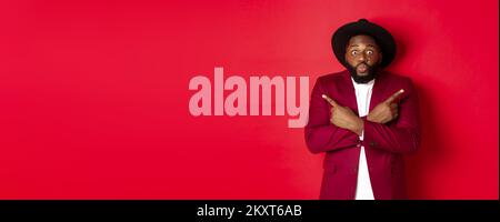 Christmas shopping and people concept. Indecisive Black man choosing between two variants, pointing fingers sideways and staring confused camera, red Stock Photo