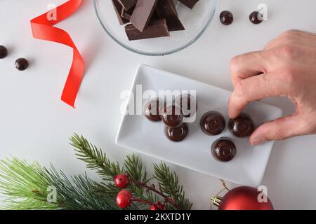 Hand picking up a chocolate bonbon from a plate on white table with more sweets and Christmas decoration. Top view. Stock Photo