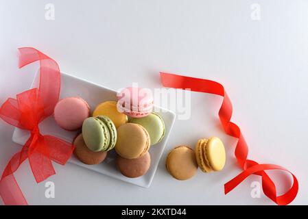 Presentation of ssortment of french macaroons of various flavors in white plate on white table with red bow. Top view. Stock Photo