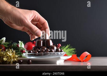 Hand picking up a chocolate bonbon from a plate on wooden table with Christmas decoration and isolated dark background. Front view. Stock Photo