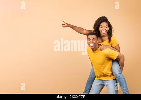 Overjoyed african american couple having fun, lady piggybacking guy and pointing finger at copy space, banner Stock Photo