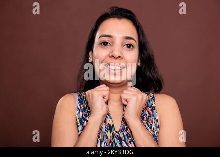 Smiling indian woman with clenched fists near cheeks medium close up portrait, cute pose. Cheerful lady touching face with hands, model with carefree expression, looking at camera Stock Photo