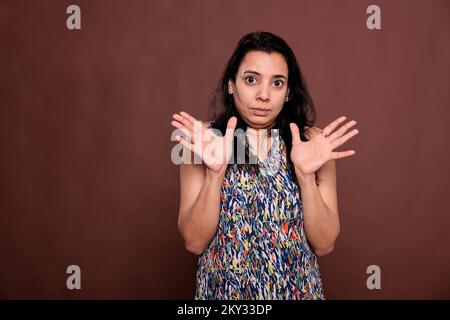 Indian woman showing no problem gesture with palms, looking at camera with confused facial expression. Young lady standing with spread arms portrait, front view studio medium shot on brown background Stock Photo