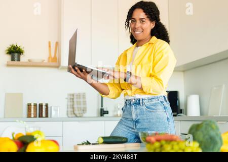 Searching for new recipes. Happy black woman holding and using laptop while cooking fresh salad in modern kitchen Stock Photo