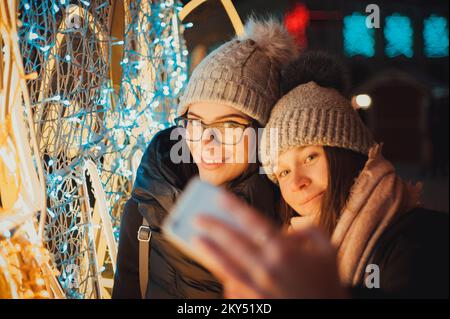 Two funny taking a selfie female friends in winter hats having fun together on the street with the background of a luminous Christmas tree outdoors Stock Photo